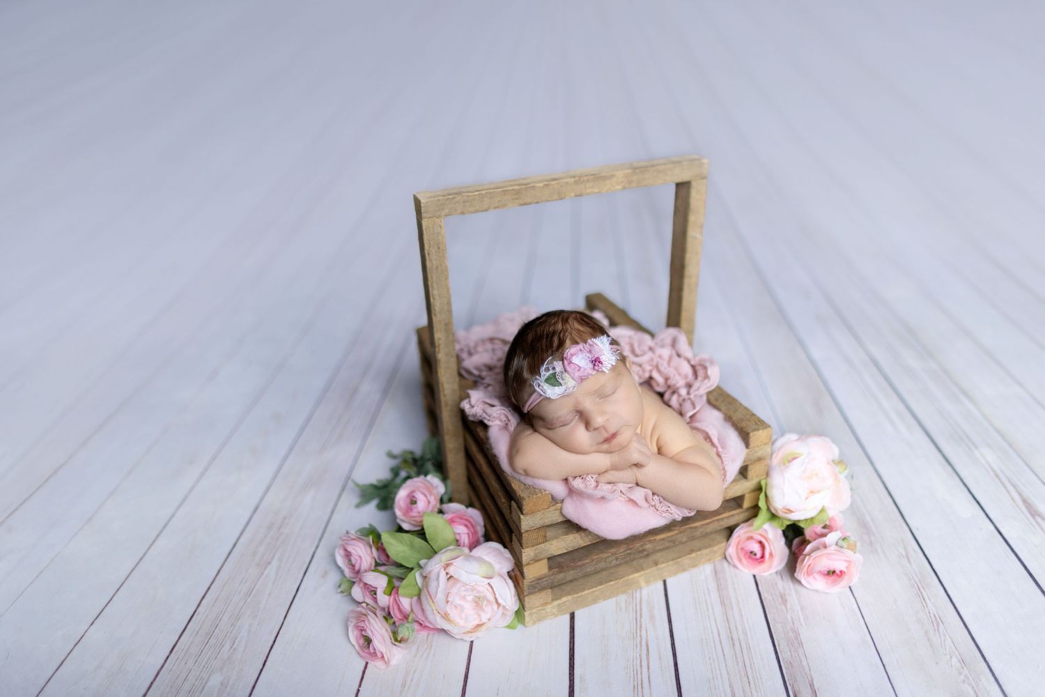 Sleeping newborn baby in pink wrap nestled in wooden crate with delicate floral decorations on white wooden floor.