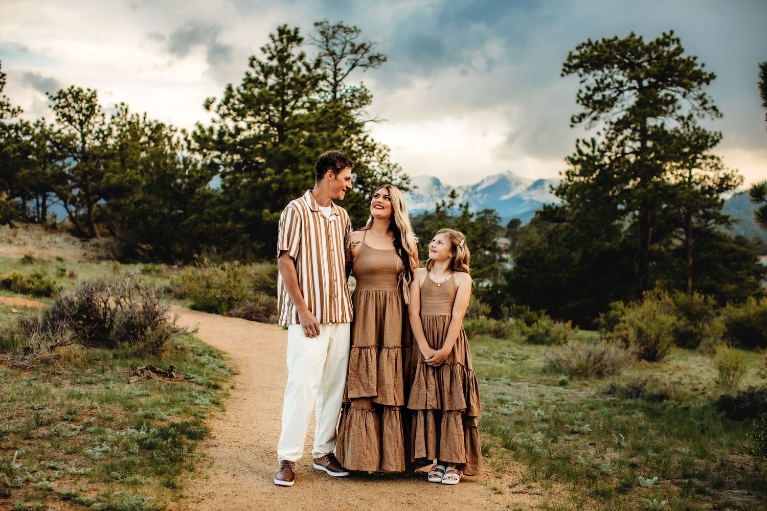 A group of people in earth-toned outfits stand together on a winding dirt path during golden hour in a natural setting.