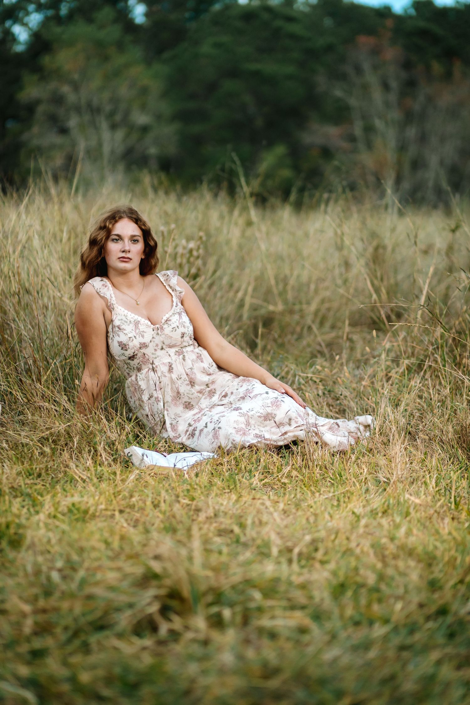 A person in a floral white dress reclines in a field of tall grass with trees in the background.