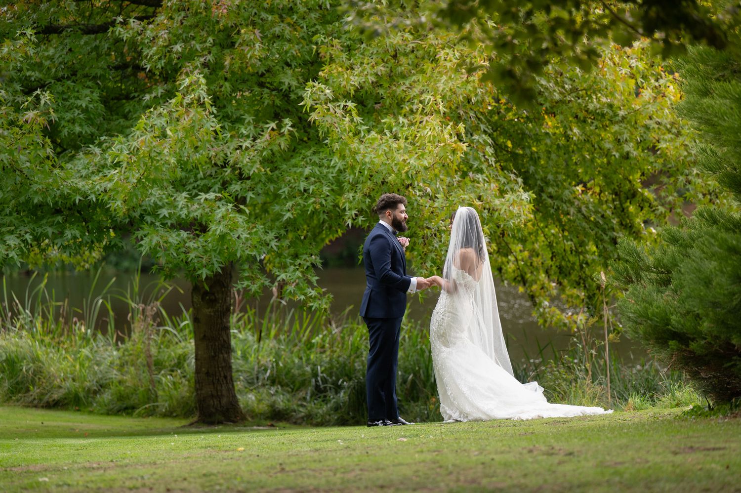 A couple in wedding attire walks through a lush green garden setting during their outdoor wedding photo session.