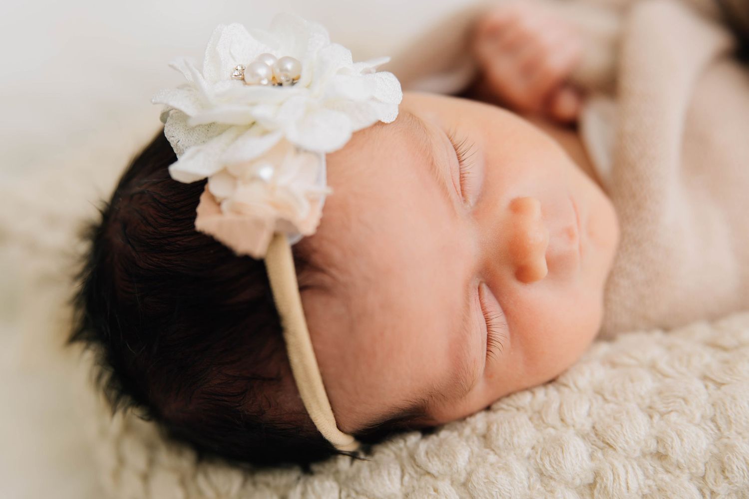 Sleeping newborn baby with dark hair wearing a delicate white flower headband on a knit blanket.