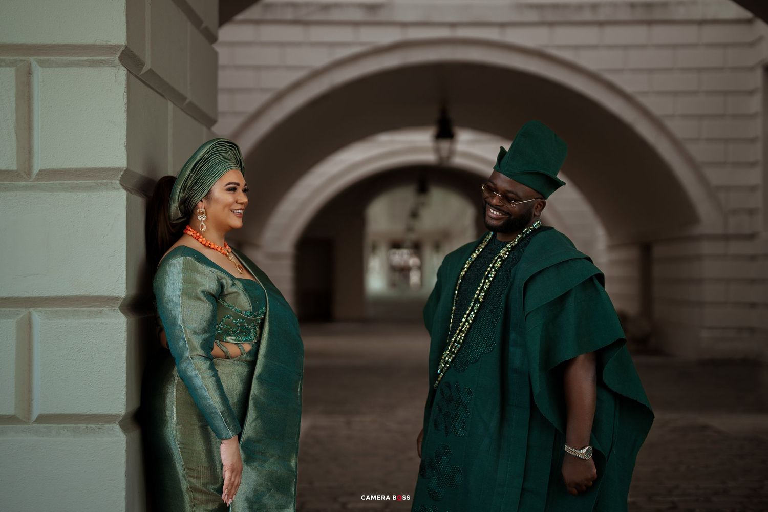 Two people dressed in emerald green attire walk hand in hand through an ornate architectural corridor.