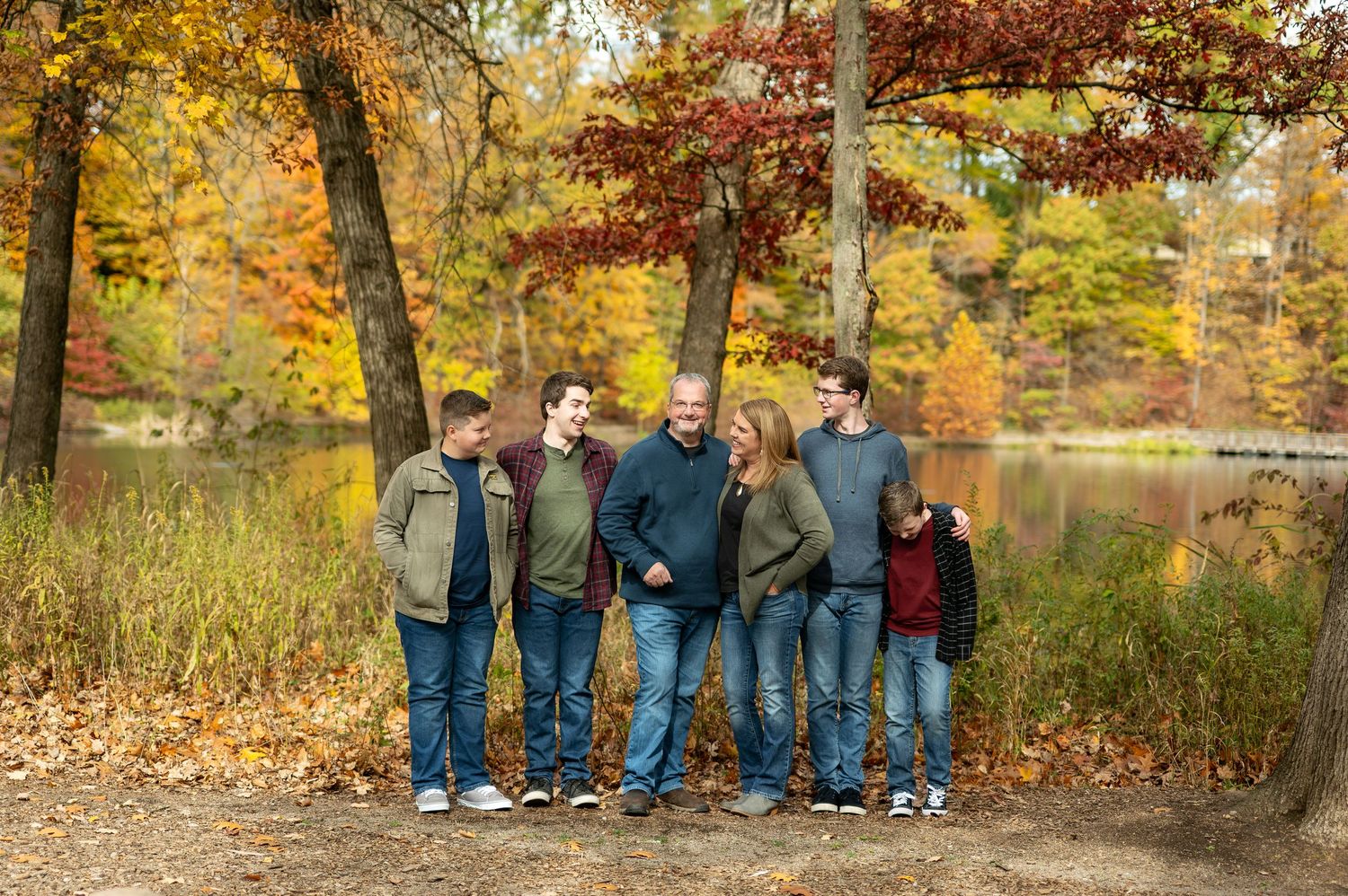 Family of six smiling together near a lake with colorful fall foliage backdrop.