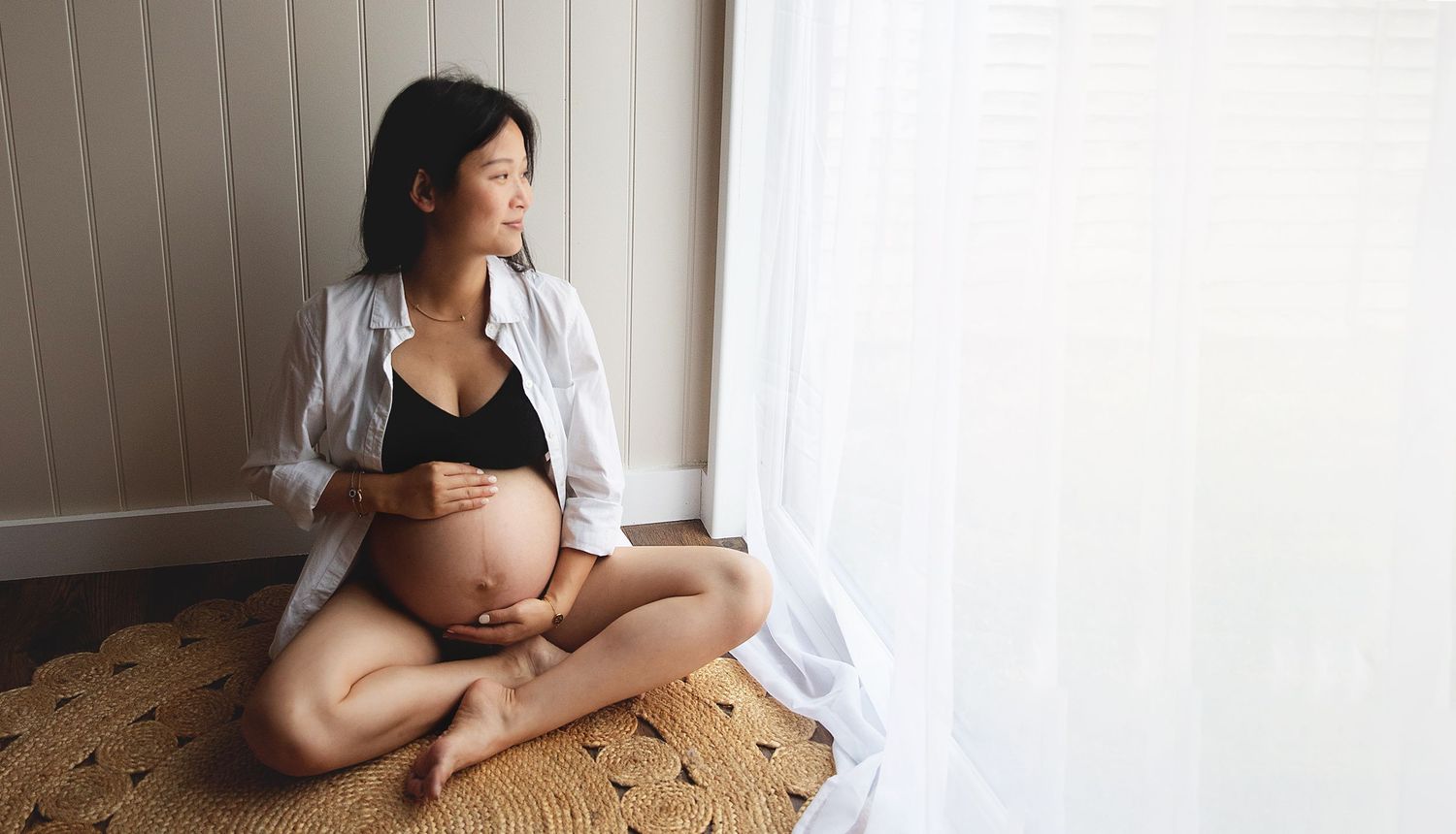 Pregnant woman sitting cross-legged by sheer curtains, cradling her baby bump in open shirt. During a maternity session in Bexley
