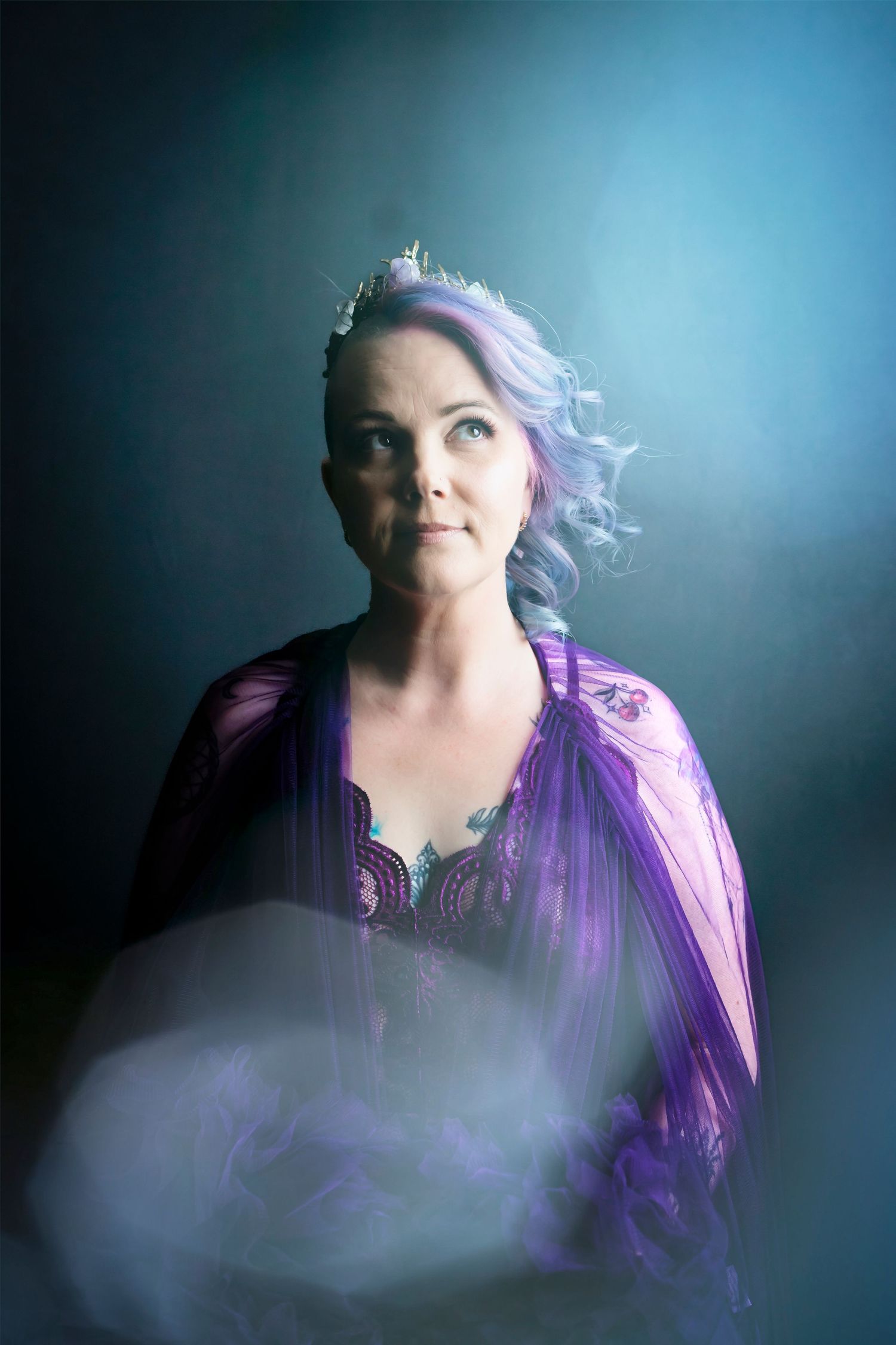 Portrait of someone in a purple gown with lavender hair against a blue backdrop with dreamy smoke effects.