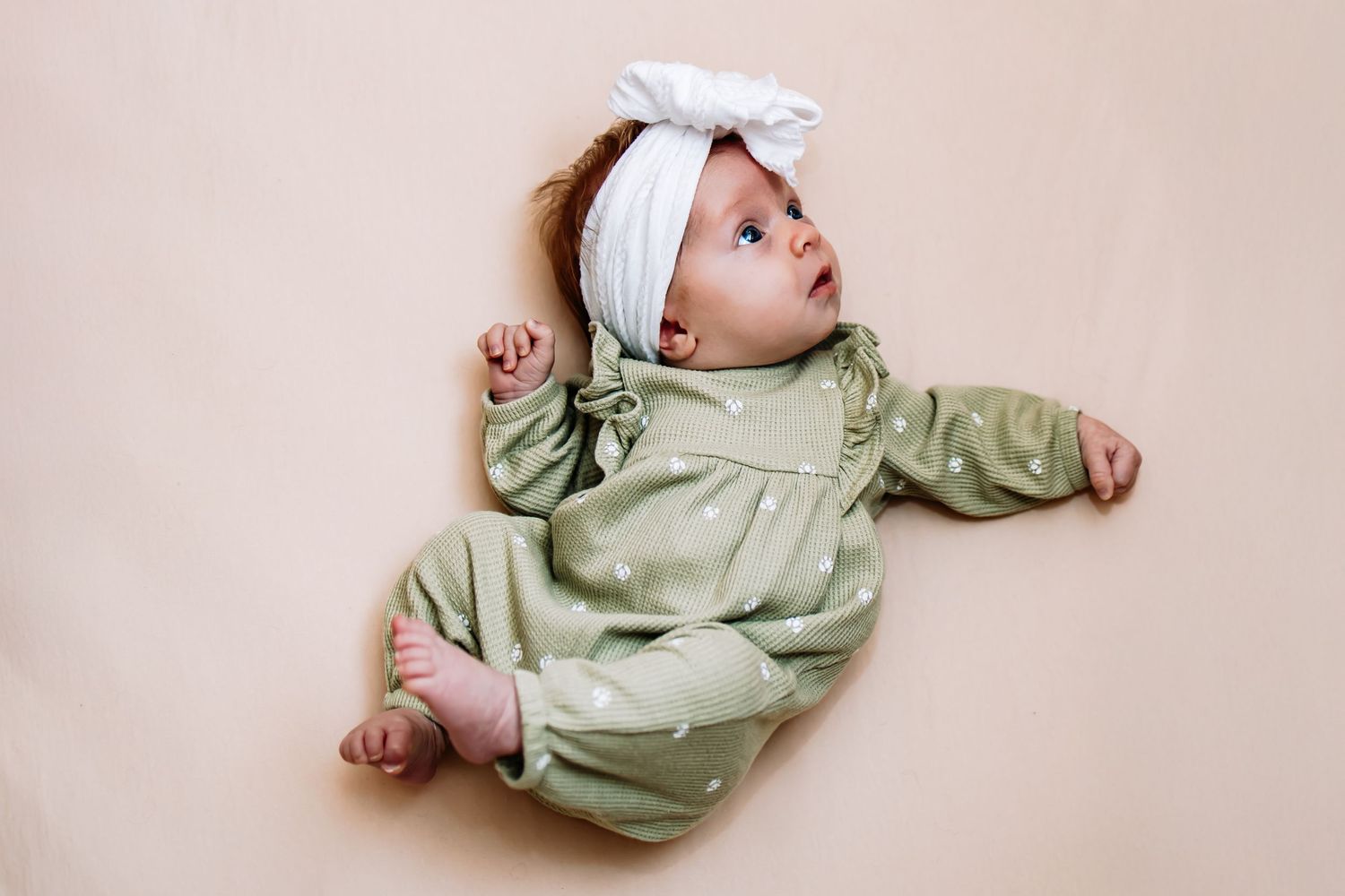 A small baby wearing a green outfit lies curled up on a neutral beige background. Lifestyle session and newborn session in Richmond, VA