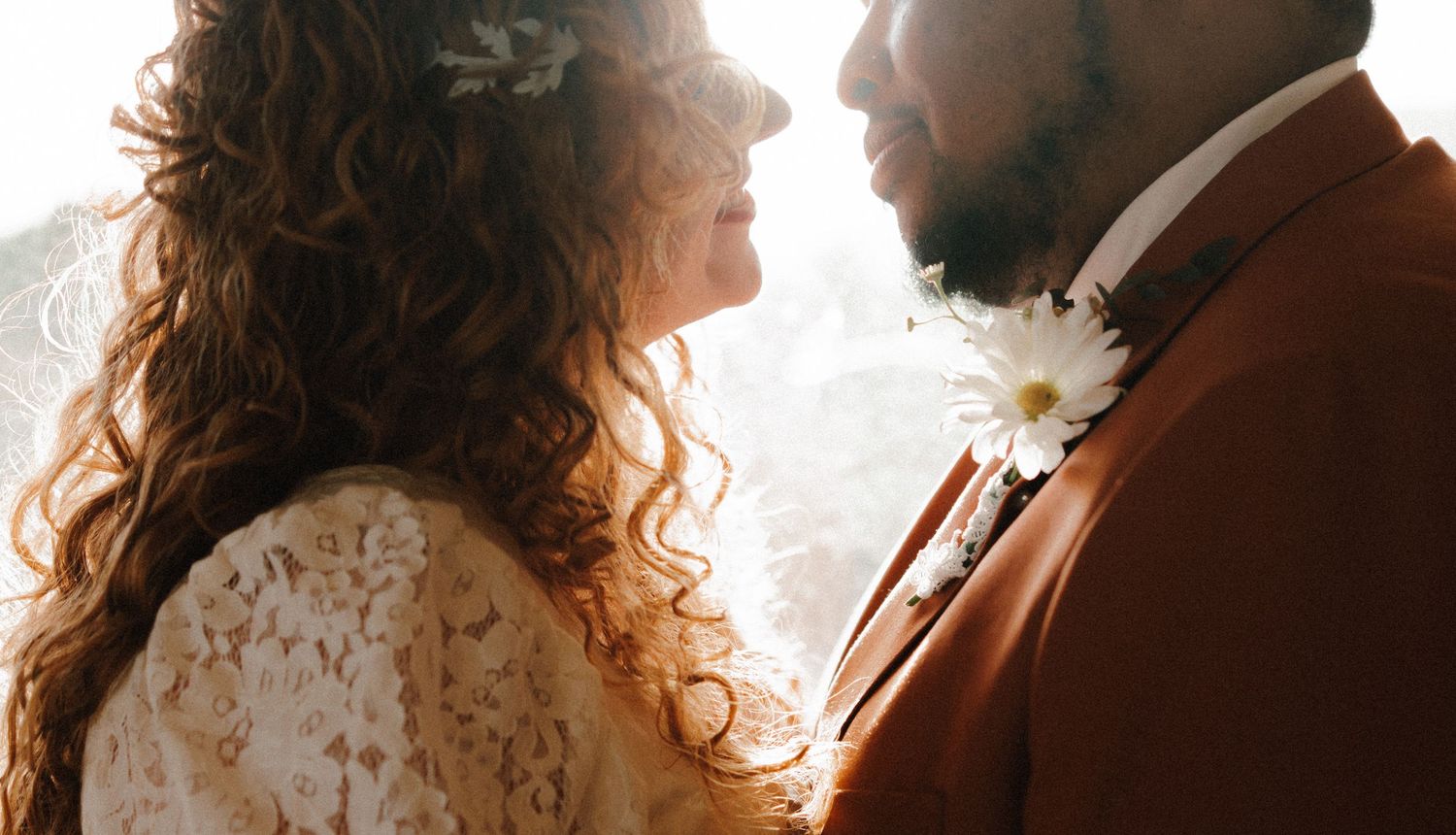 Sunlit wedding photo captures intimate moment between newlyweds, with lace gown and brown suit details.