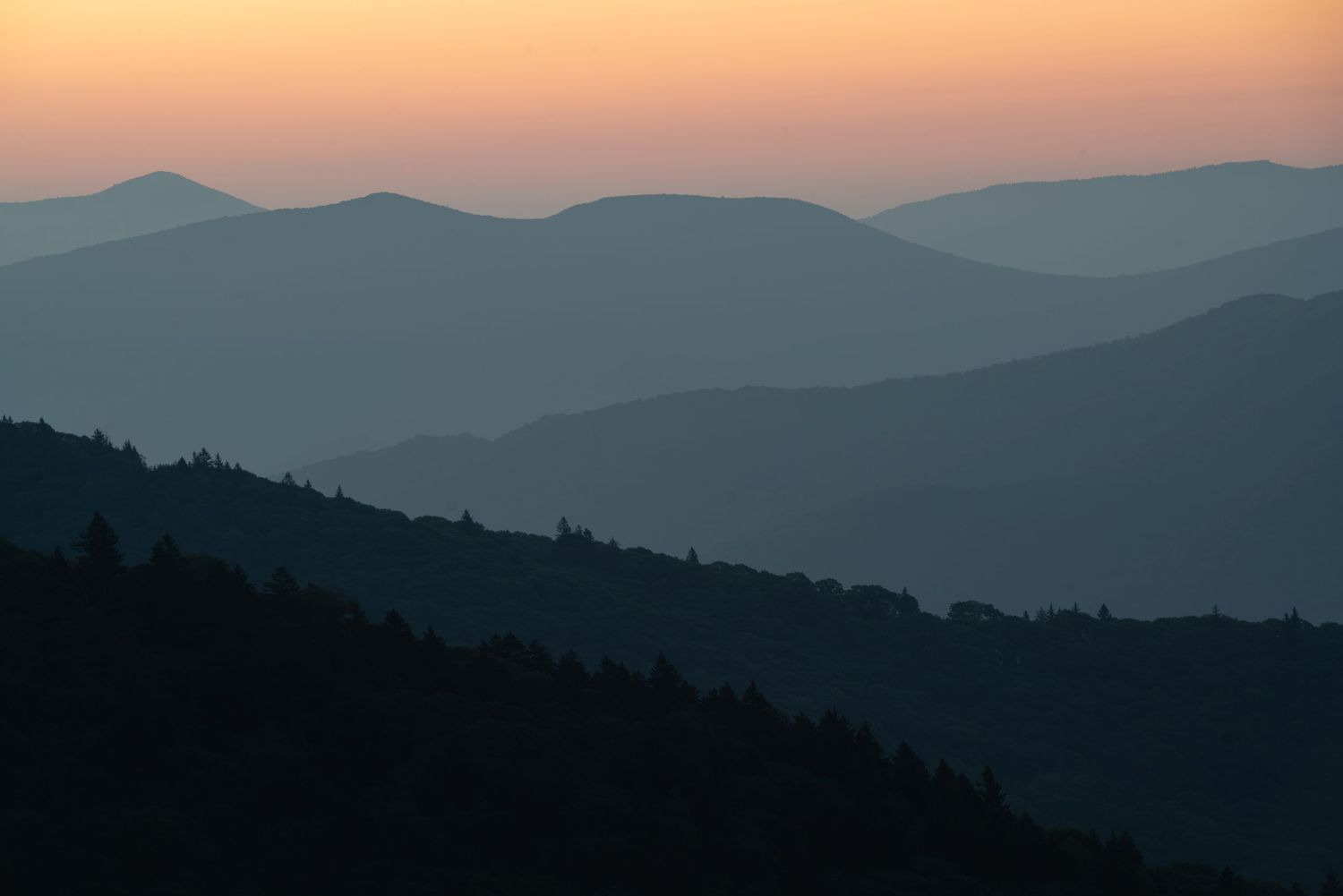Layered blue mountain silhouettes fade into a warm orange sunset sky in a serene landscape panorama.