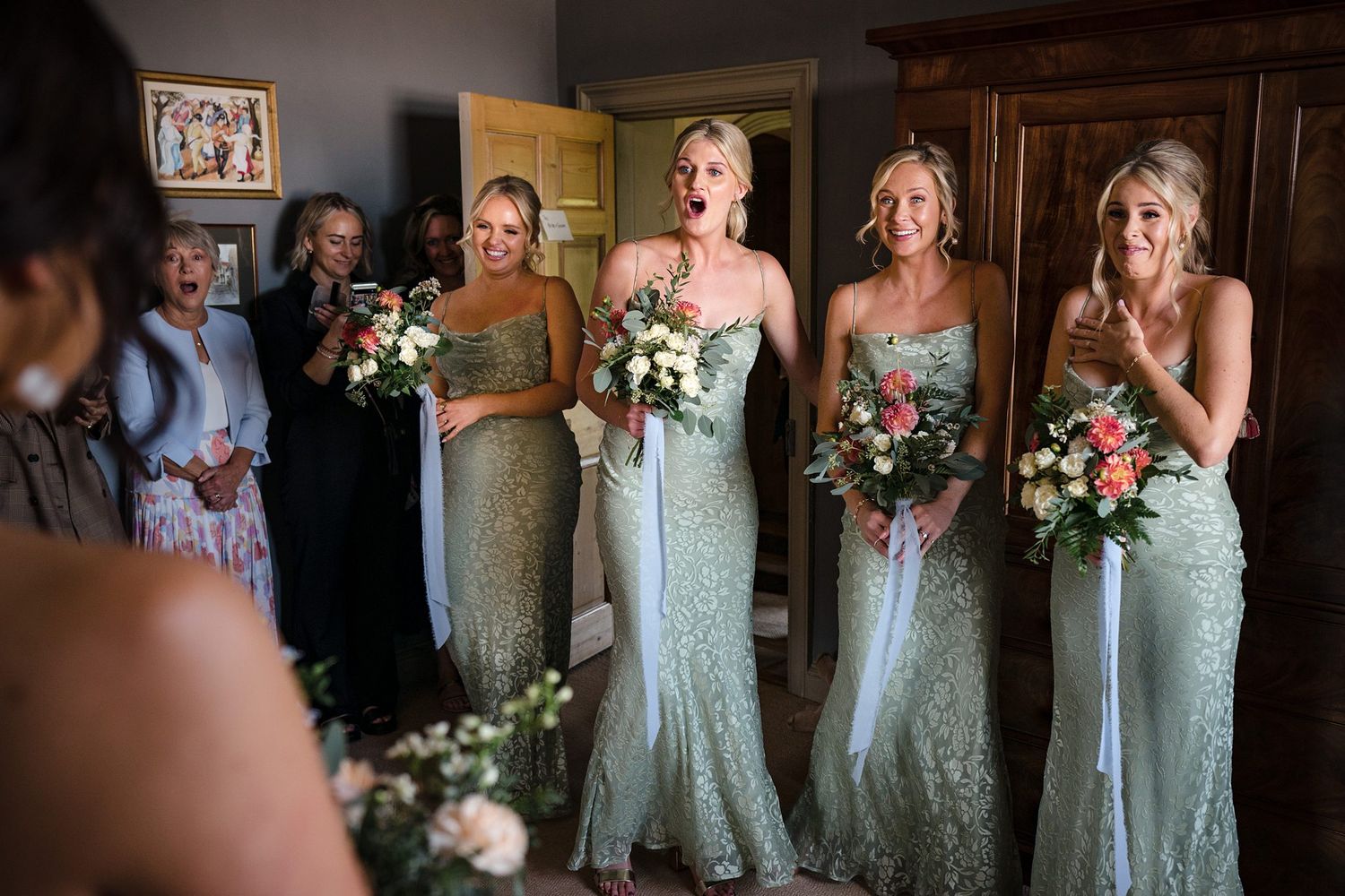 Bridesmaids in sage green lace gowns holding coral and white bouquets smile at bride during wedding preparation.