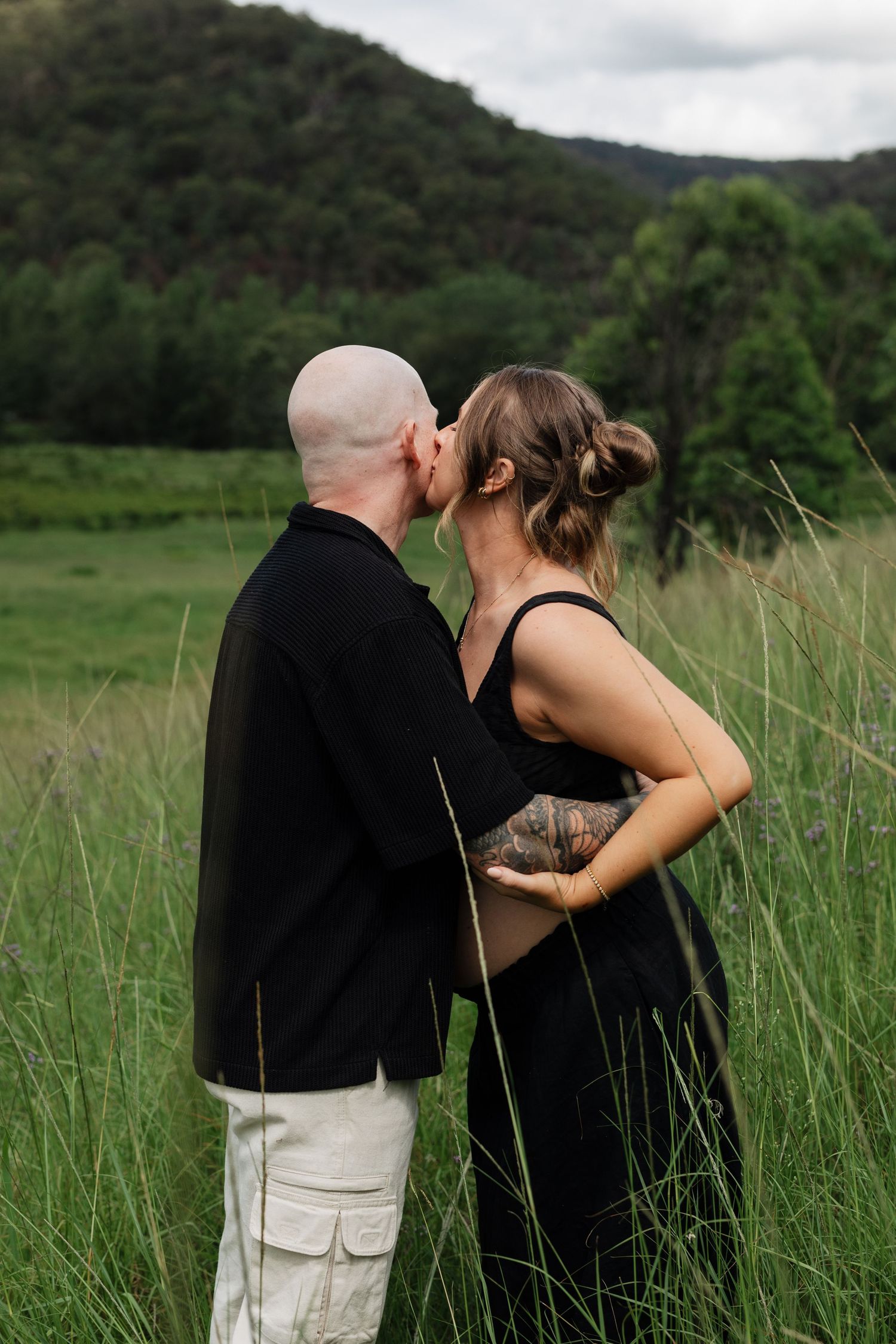 A couple embraces in a serene meadow with tall grass and mountains in the background.