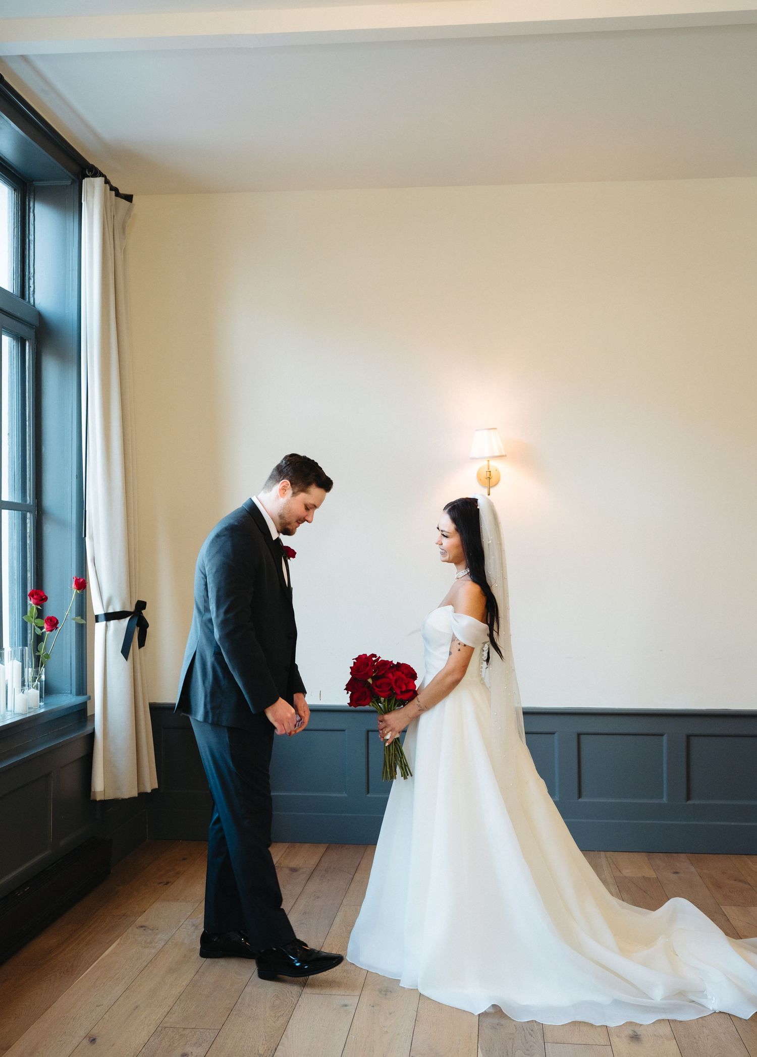 A couple in formal wedding attire share a tender moment in a bright minimalist room with wooden floors.