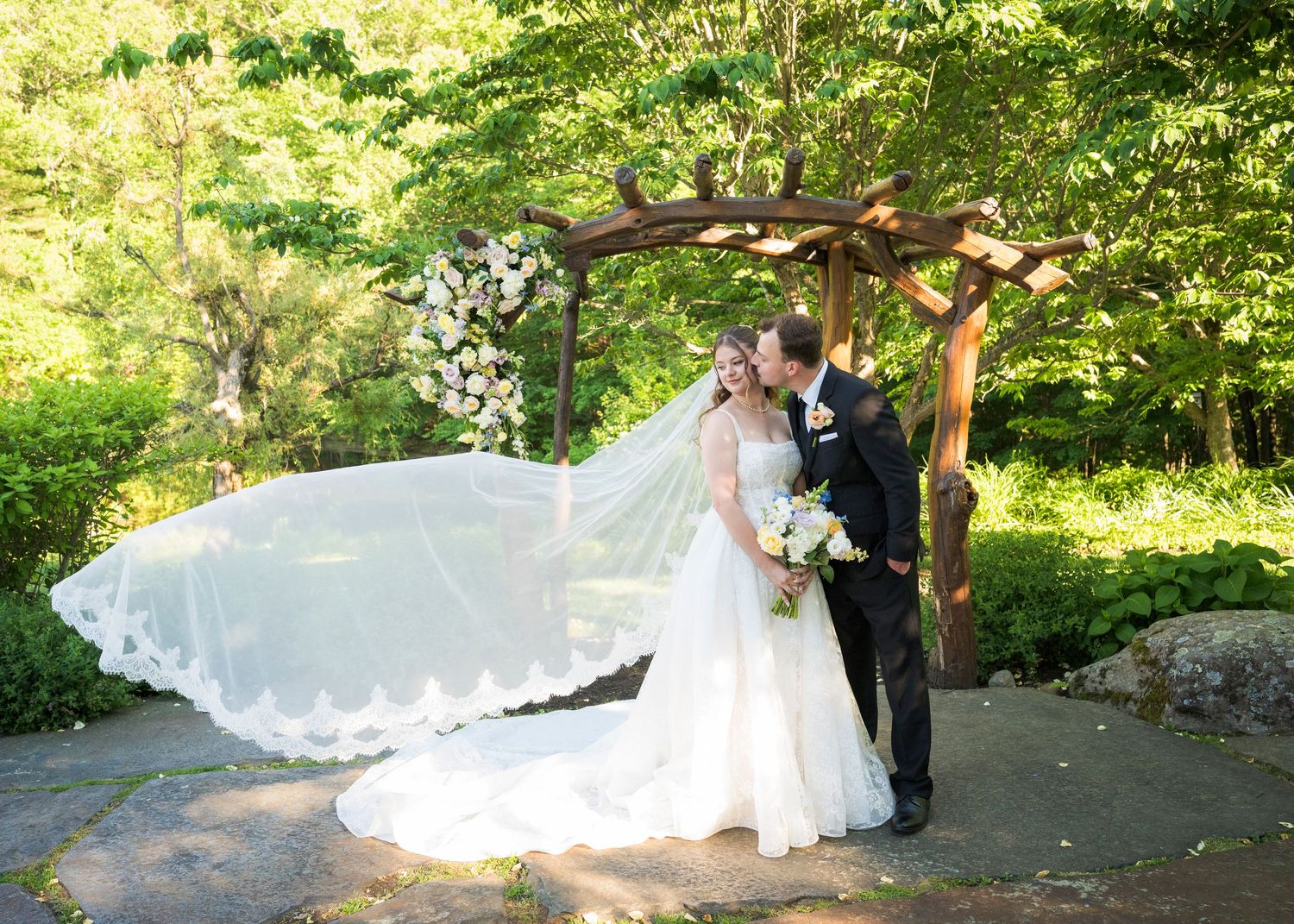 Bride and groom kissing under rustic wooden arch with flowing veil in lush green outdoor wedding ceremony.
