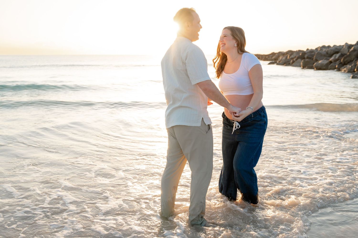Romantic couple embraces on sandy beach at sunset with ocean waves and golden light in the background.