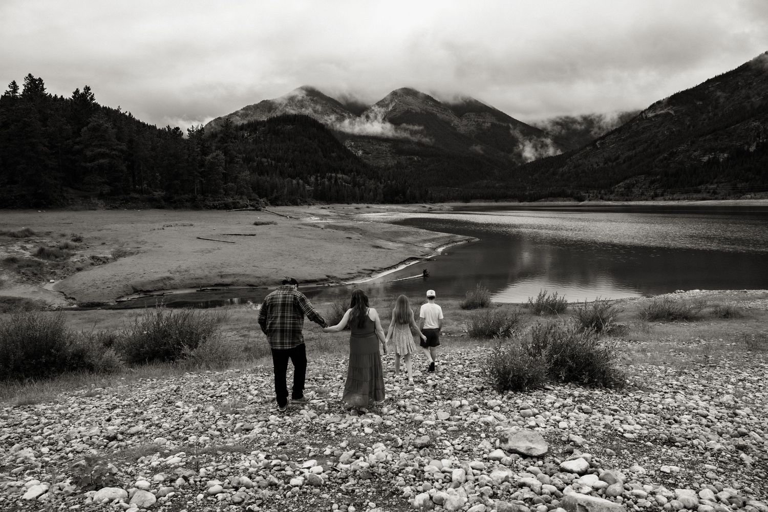Black and white photo of three people holding hands facing a mountain lake under cloudy skies.