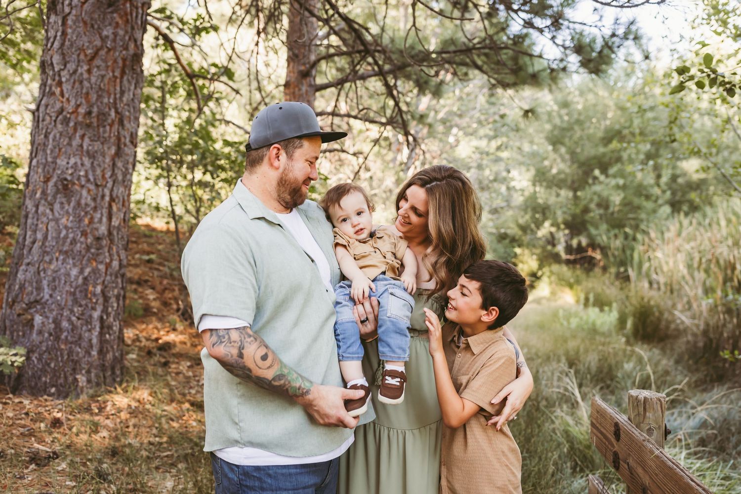 Family of four smiling outdoors among pine trees, parents holding young children in a warm natural setting.
