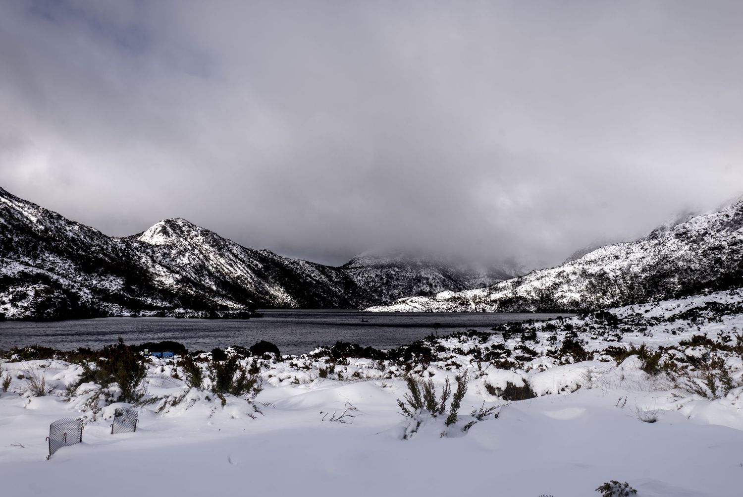 Snow-covered mountain valley with a frozen lake, dark storm clouds, and winter landscape in a remote wilderness setting.