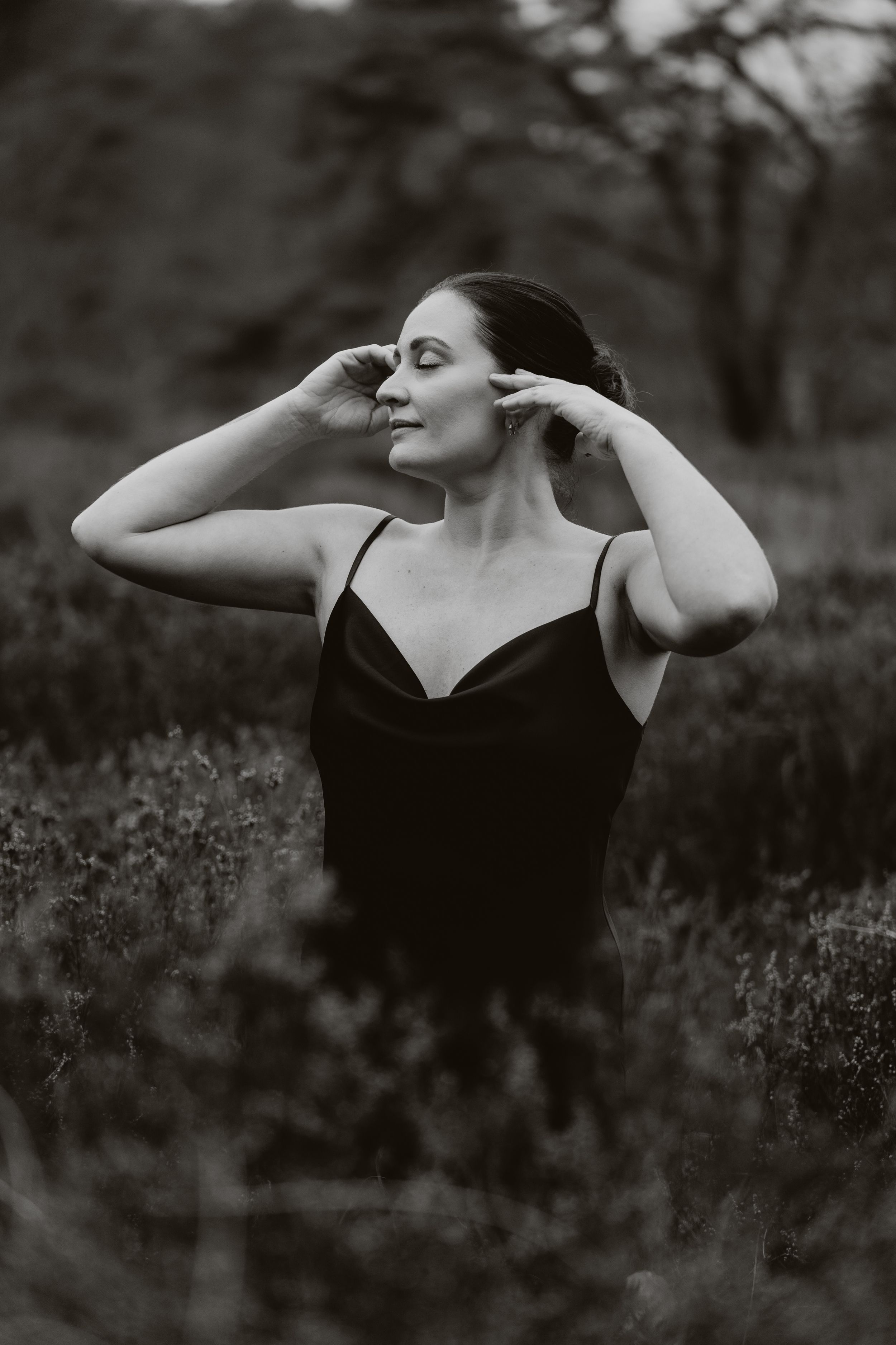 Black and white portrait of a woman in a dark slip dress standing in a field, hands raised to her hair, eyes closed.