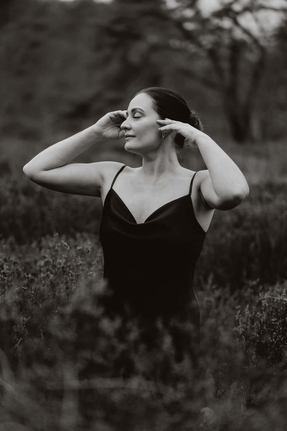 Black and white portrait of a woman in a dark slip dress standing in a field, hands raised to her hair, eyes closed.
