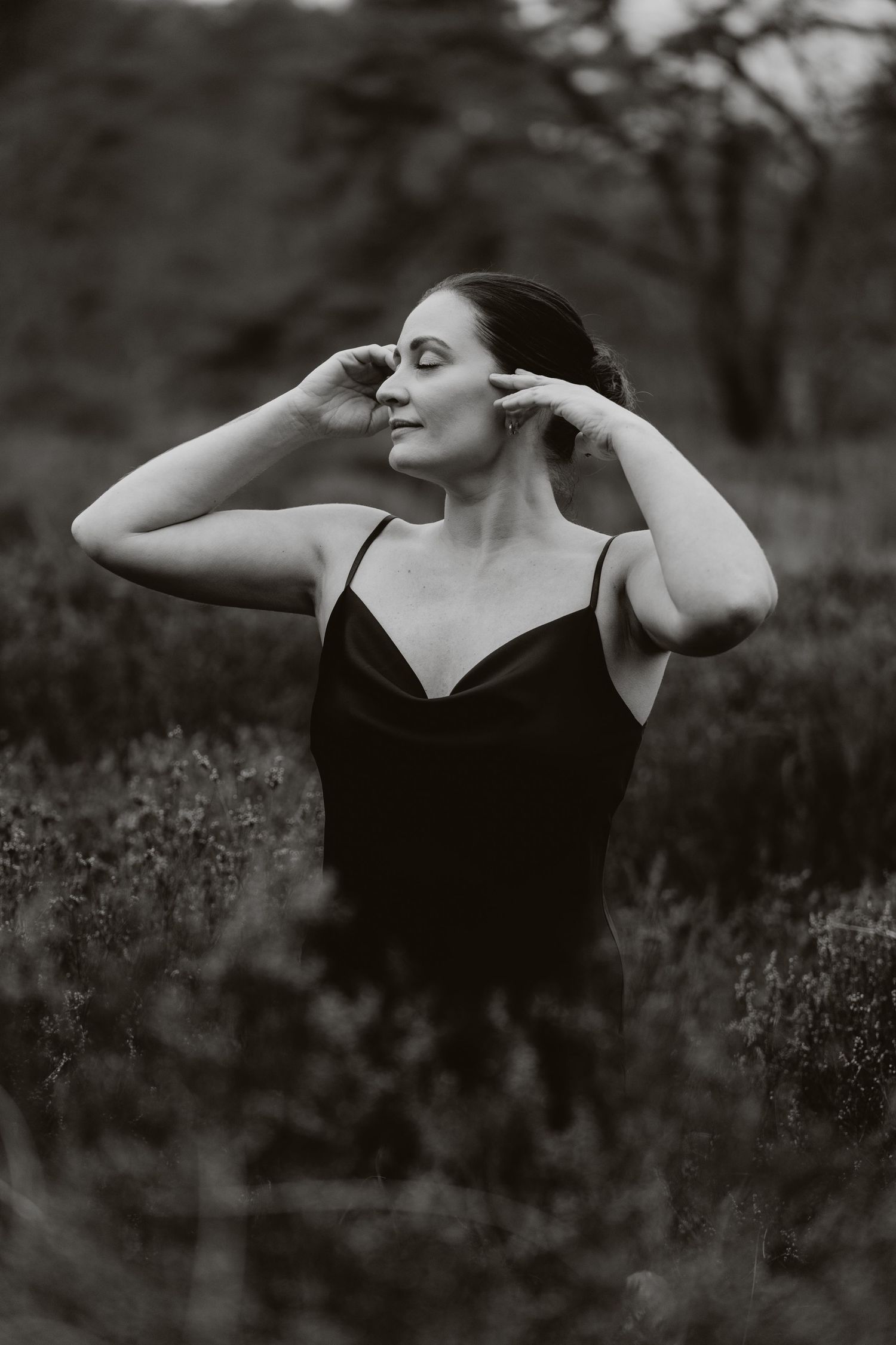 Black and white portrait of a woman in a dark slip dress standing in a field, hands raised to her hair, eyes closed.