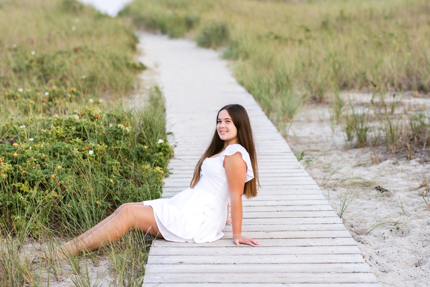 Girl with long dark hair wears white dress for senior photos sitting on beach boardwalk with coastal grass. North Shore senior photographer