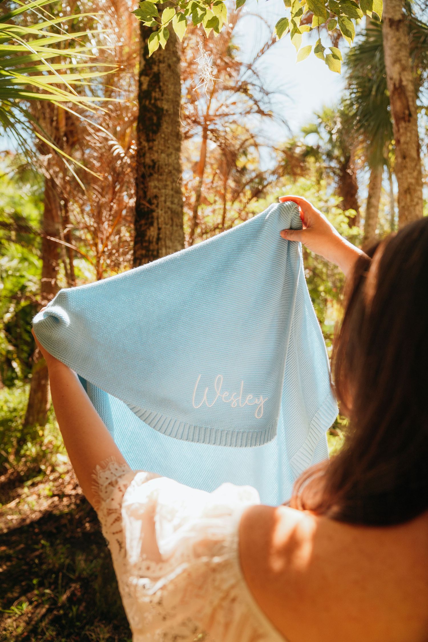 A light blue fabric is held up against sunlight in a forest setting, creating a dreamy ethereal effect.