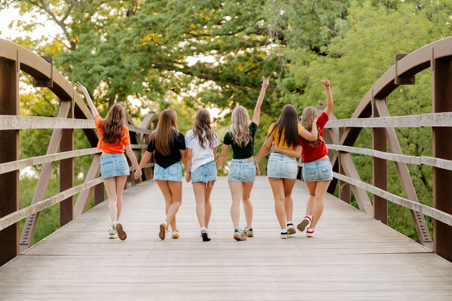 Five young women in white outfits hold college pennants on a tree-lined path celebrating their university commitments.