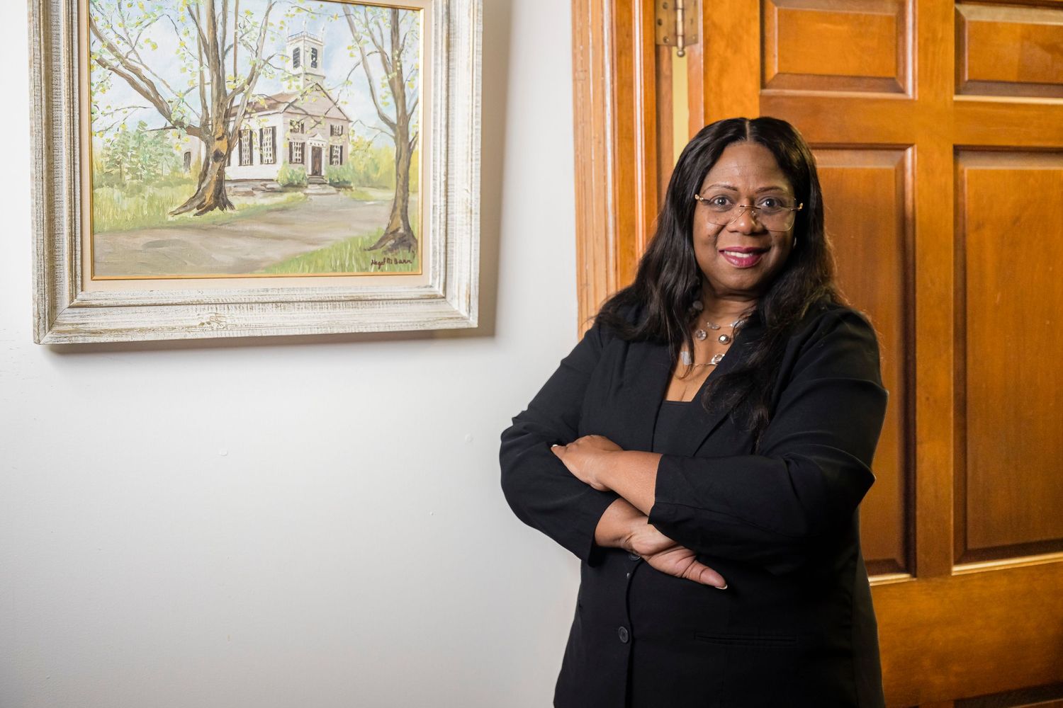 Professional woman in black blazer stands confidently with arms crossed beside a painting near a wooden door.