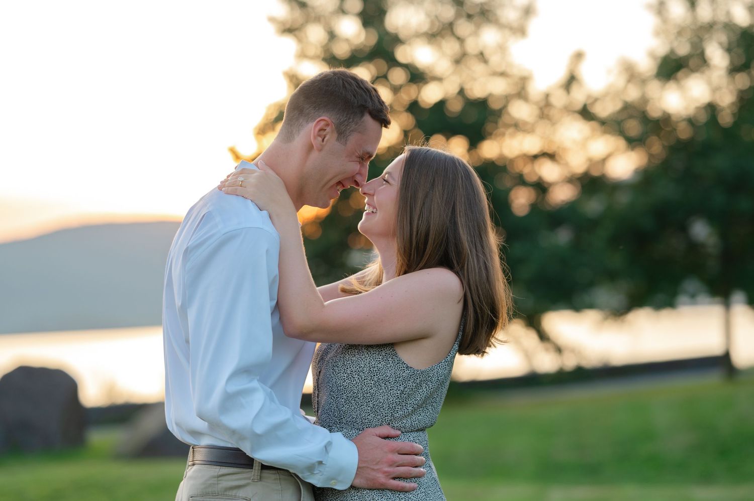 Couple embracing and smiling at sunset in a green park with trees and water in the background in Peekskill, NY during a proposal session.