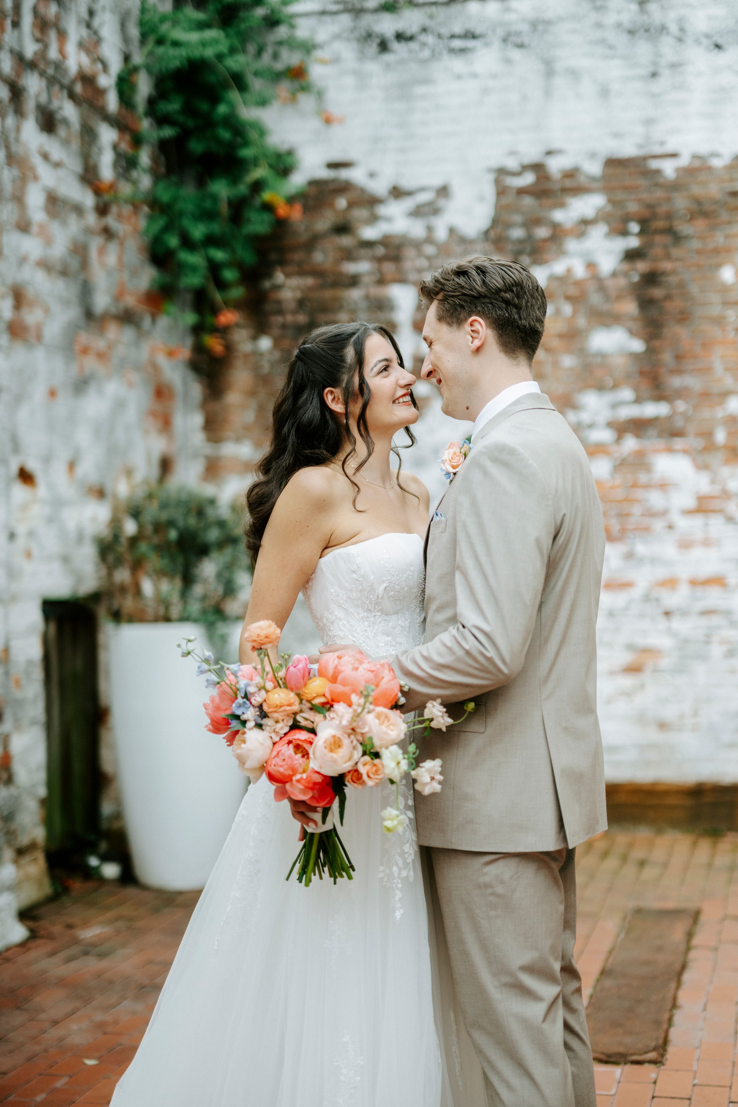 Bride and groom share a romantic moment outdoors, bride holding a vibrant coral and pink bouquet against a rustic brick wall.
