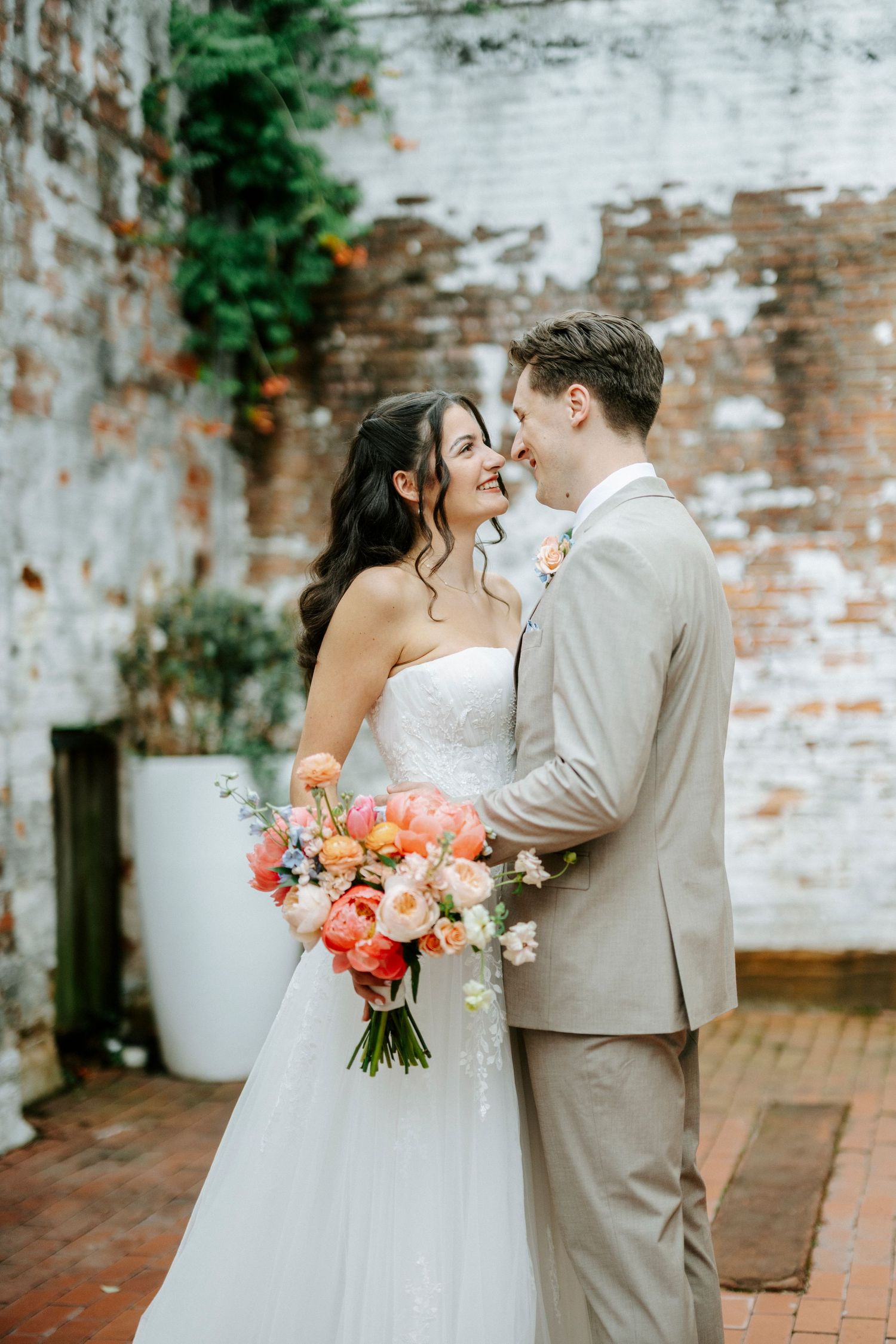 Bride and groom share a romantic moment outdoors, bride holding a vibrant coral and pink bouquet against a rustic brick wall.
