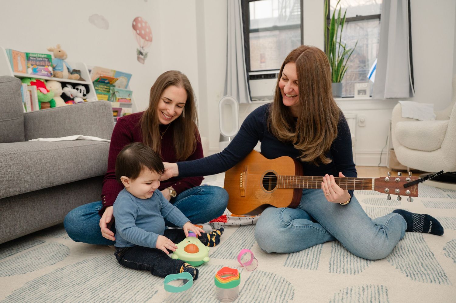 Two women and a toddler play on a carpet, one strumming a guitar during a fun indoor music session.