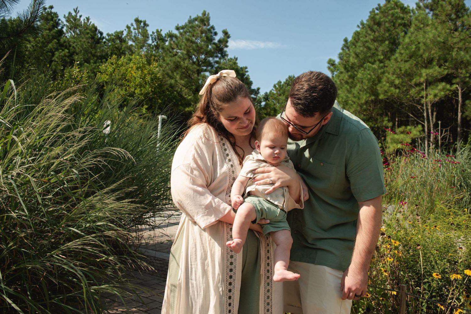 A young family with a baby poses outdoors among lush greenery and wildflowers on a sunny day.