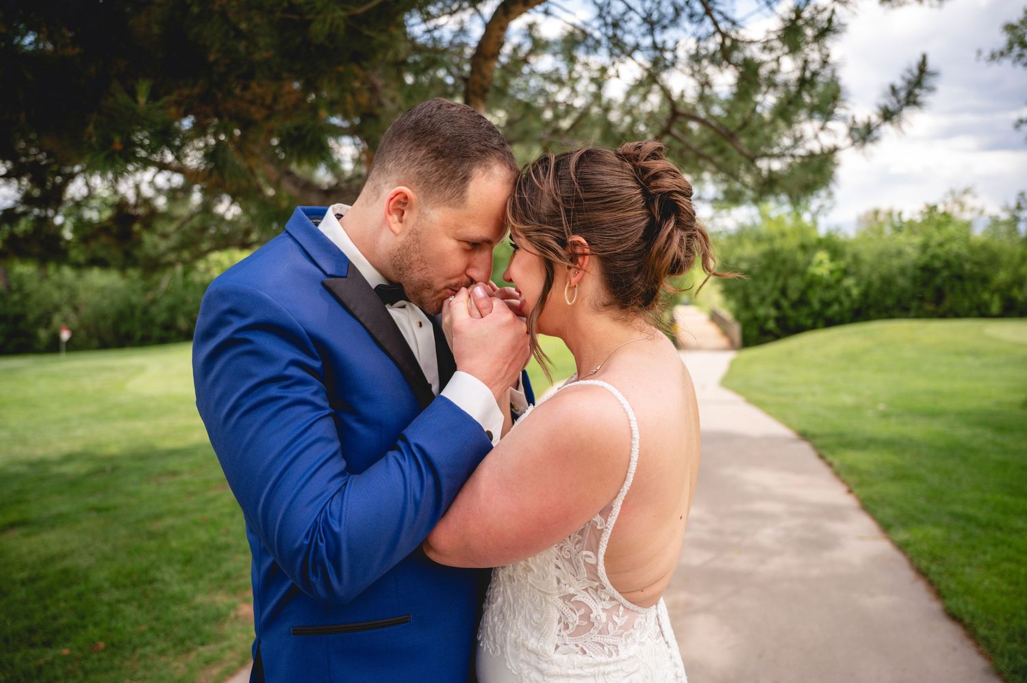 Groom in blue suit kisses bride's hands tenderly outdoors on their wedding day.