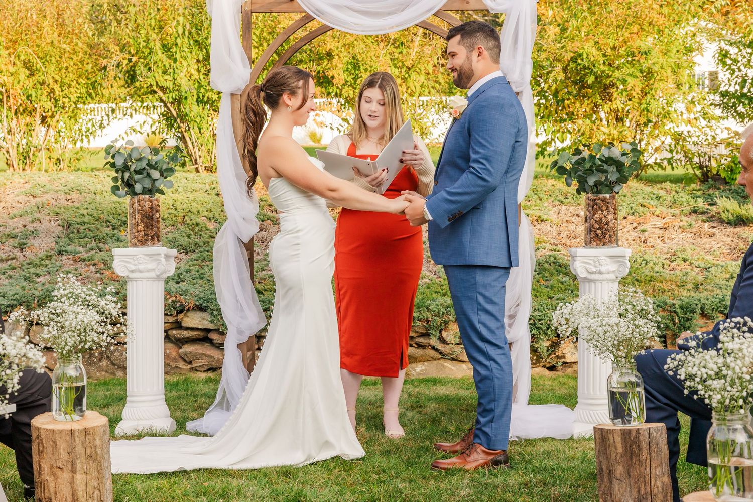 Outdoor wedding ceremony with bride and groom exchanging vows before officiant in red dress, framed by white arch and floral pillars.