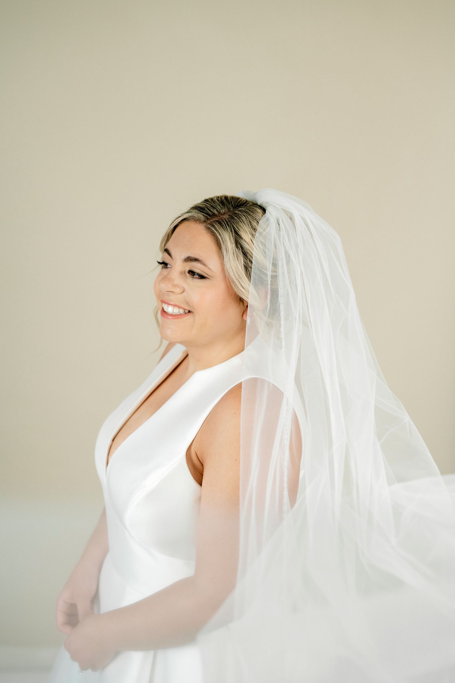 Smiling bride in a white sleeveless gown and flowing veil poses elegantly against a soft neutral background.