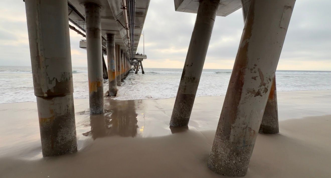 View underneath a beach pier with sand and ocean water visible between weathered concrete support pillars.