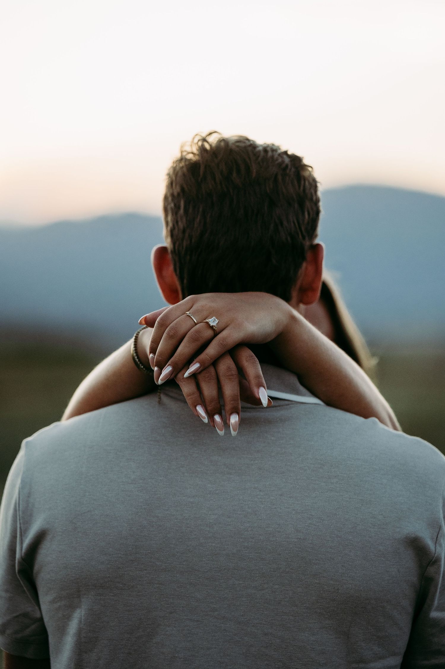 A woman with manicured nails and an engagement ring embraces a man from behind at sunset with mountains in the background.