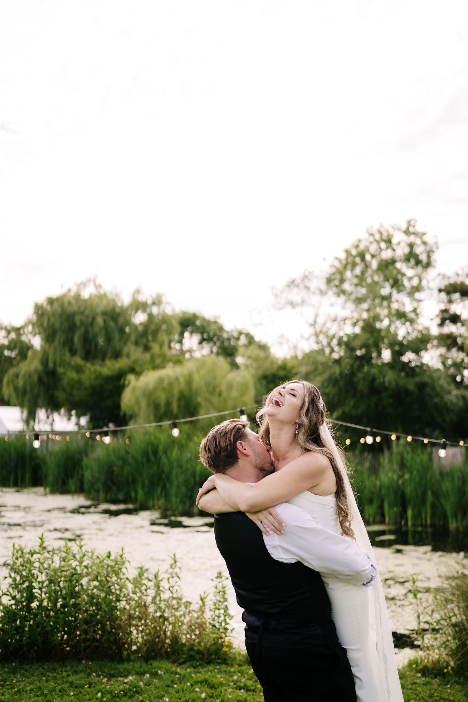 Joyful bride laughing as groom lifts her in a romantic outdoor wedding photo by a pond with willow trees. Couple Shot's with Stu Tucker