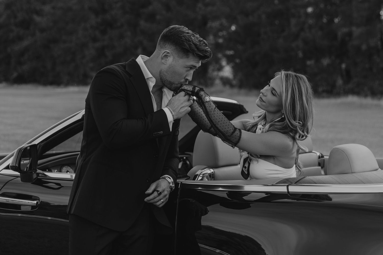 Black and white photo of an elegant couple in formal wear leaning against a convertible car outdoors.