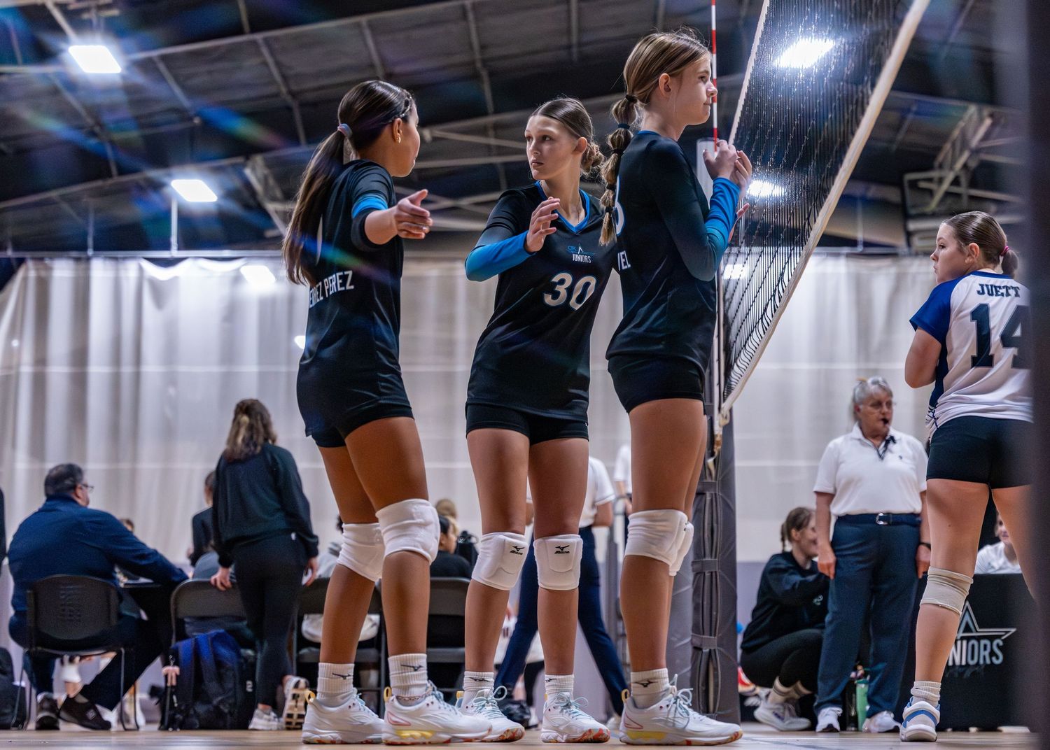 Three volleyball players in black uniforms huddle together on the court during an indoor volleyball match.