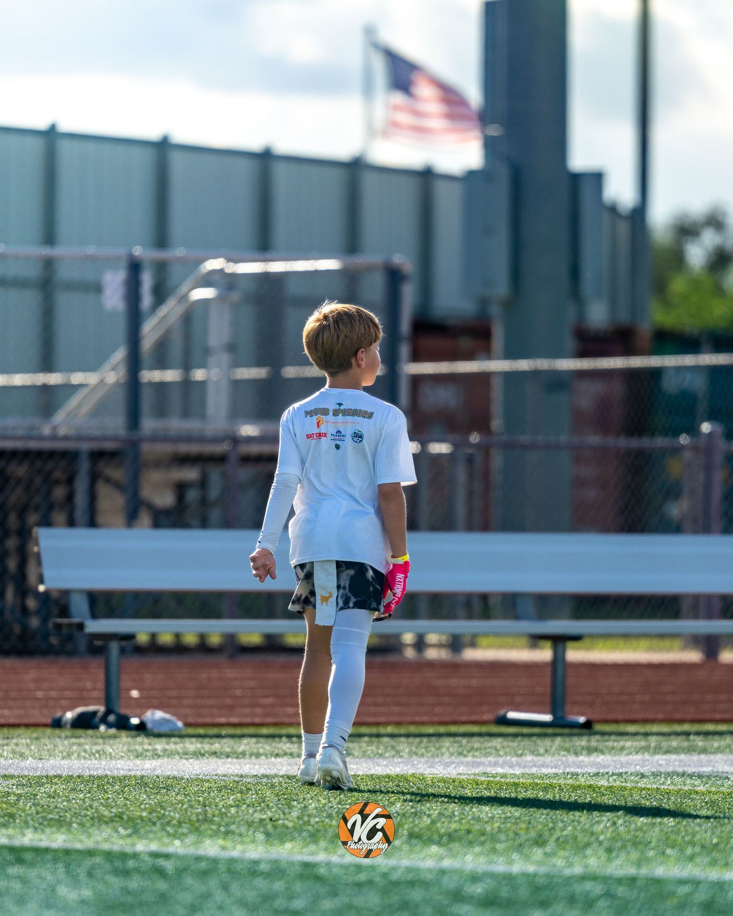 Young soccer player in white jersey stands on turf field with ball, American flag visible in background.