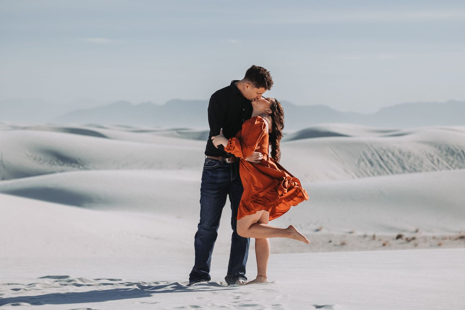 Couple sharing a romantic embrace on white sand dunes, woman in rust dress lifting her foot, misty mountains behind.