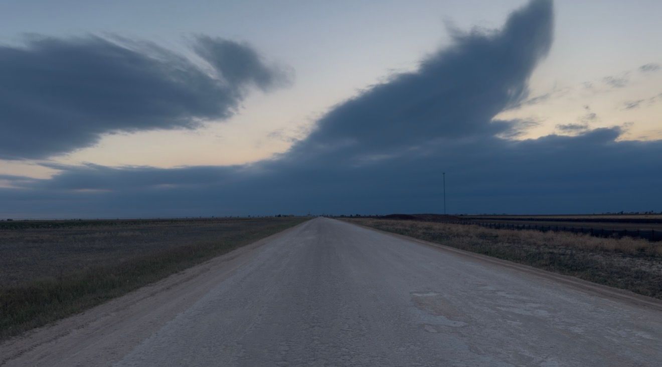 A straight rural road stretches into the horizon under dramatic storm clouds at dusk over flat open plains.
