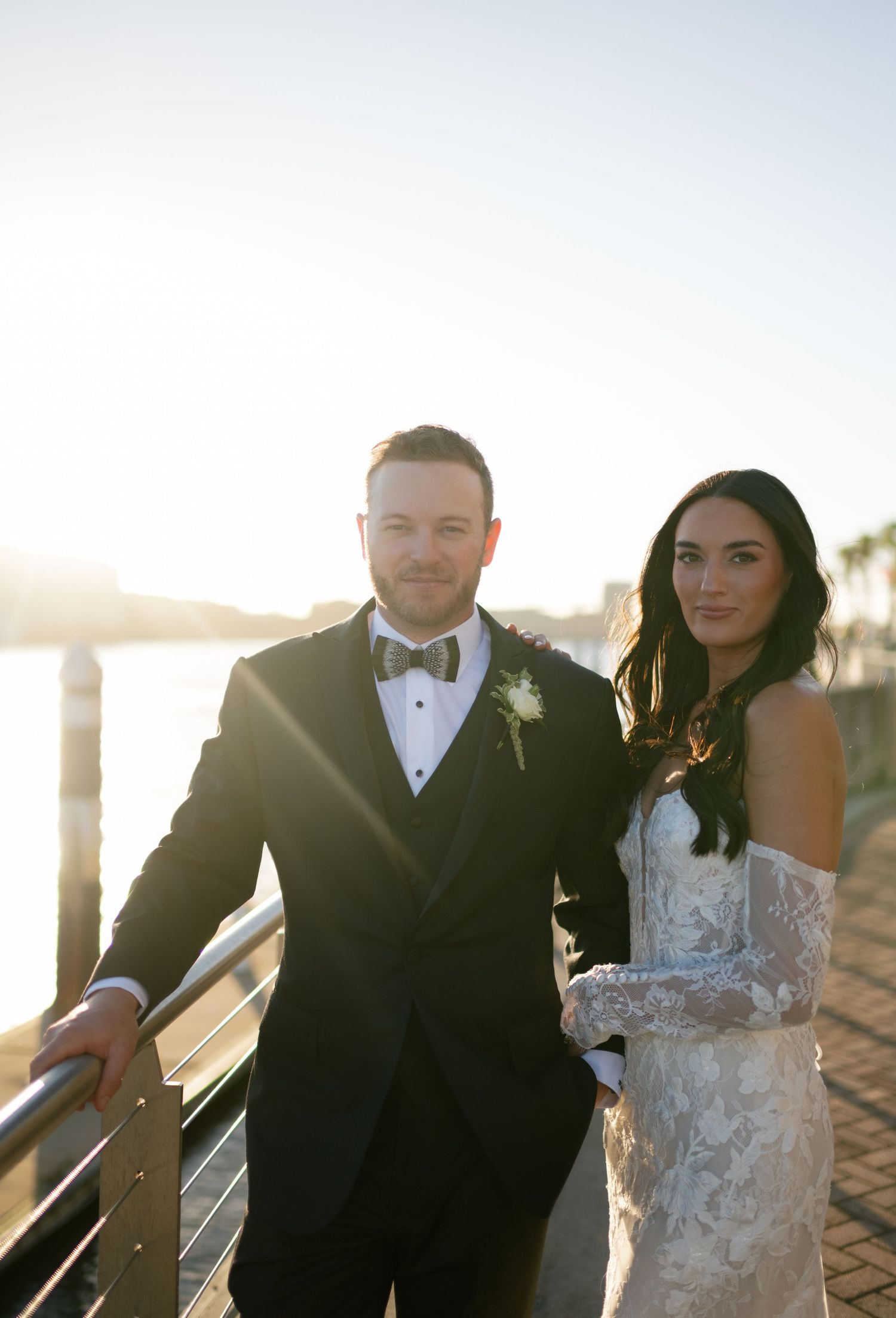 A couple in formal wedding attire poses together outdoors at sunset against a scenic backdrop.