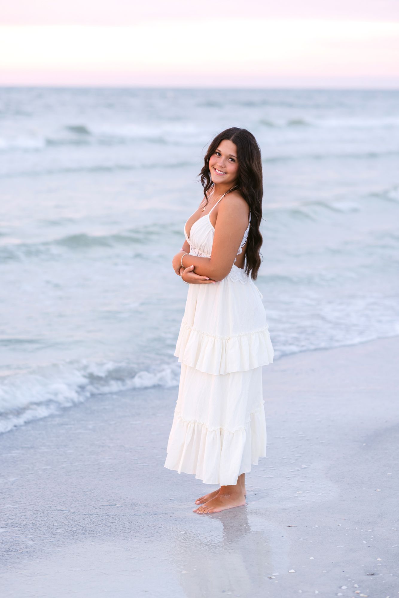 Dark, long haired girl wears a white dress and poses standing for senior photos at sunset at Indian Rocks Beach, Clearwater Florida.