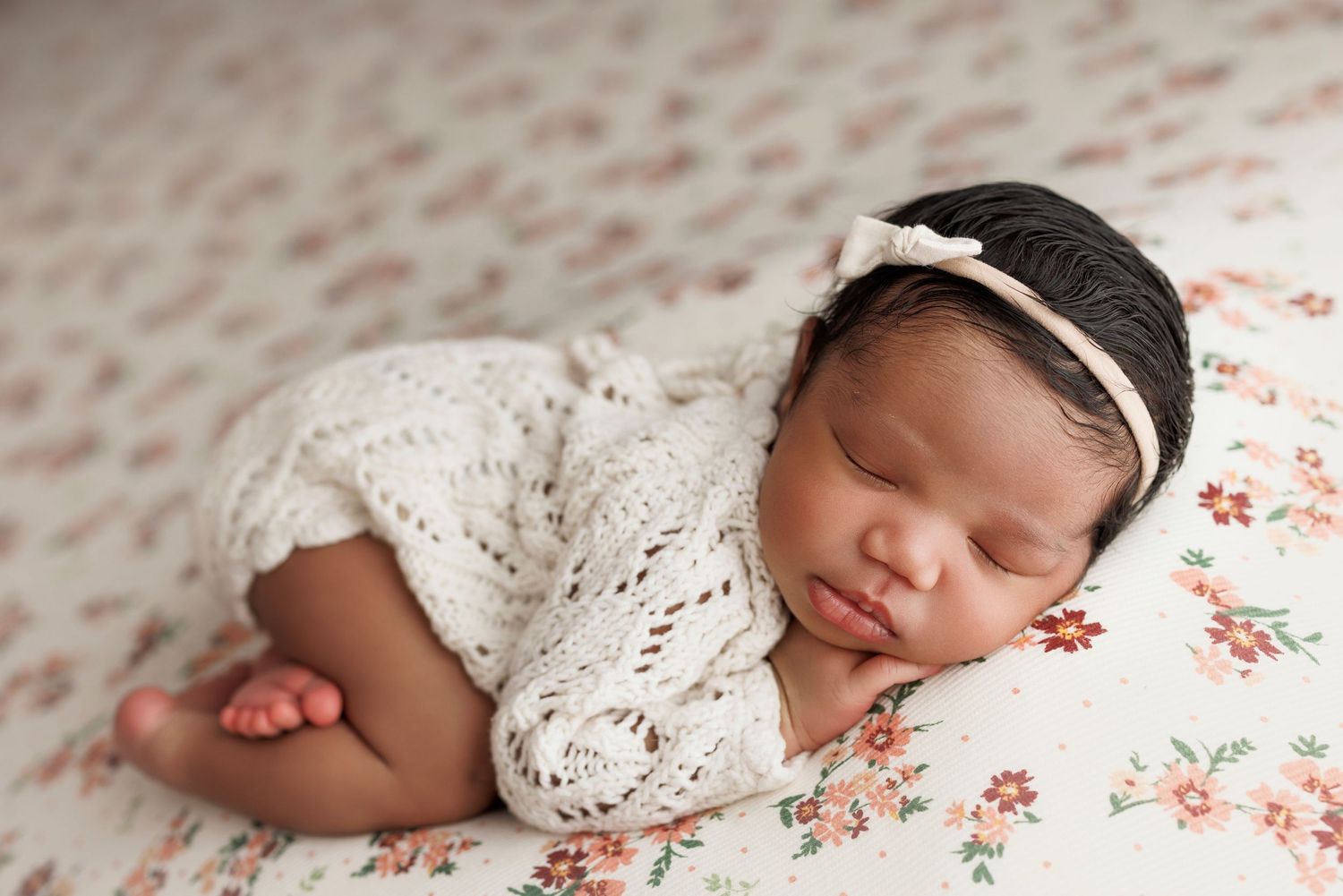 Sleeping newborn baby in white lace romper with bow headband on floral blanket.