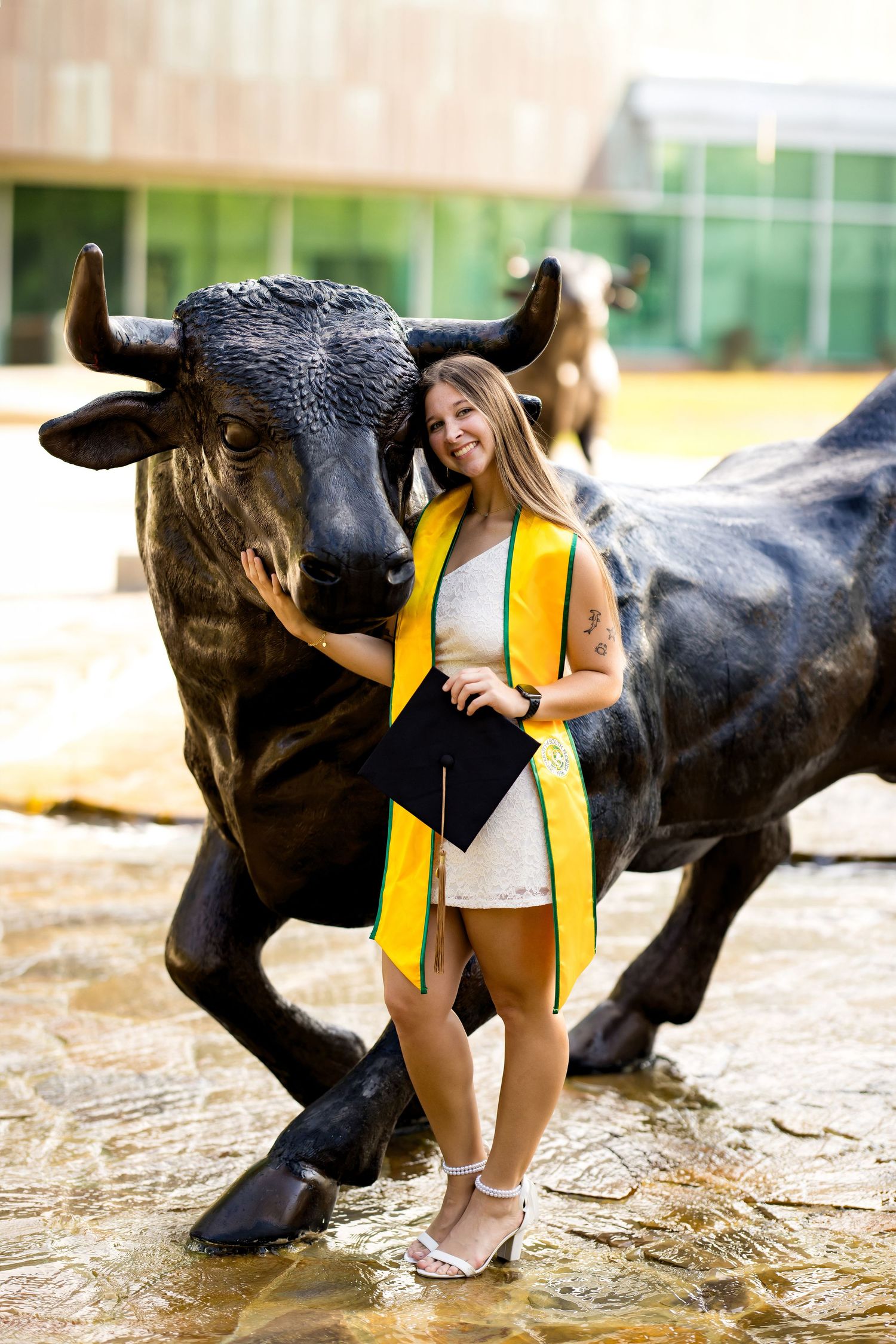 Smiling female graduate in white dress with gold sash holds cap beside bronze bull statue on campus.