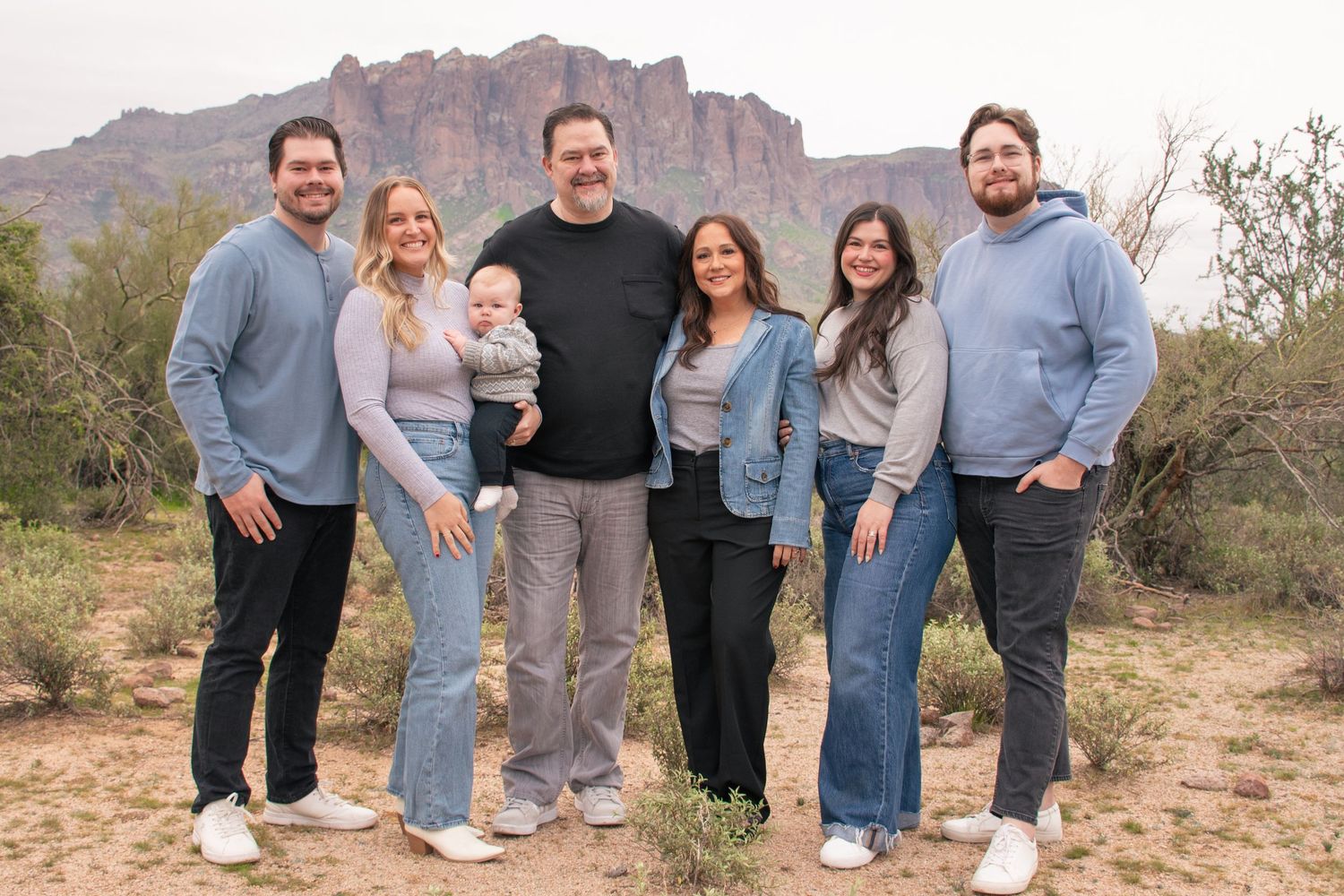 A group of people in coordinated blue and denim outfits pose together against a desert mountain landscape background.