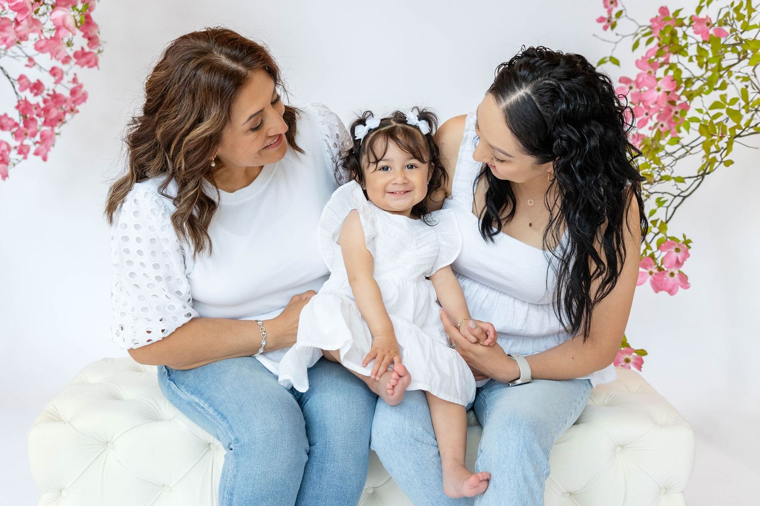 Two women in white tops and jeans sitting with a toddler girl in white dress surrounded by pink floral backdrop.
