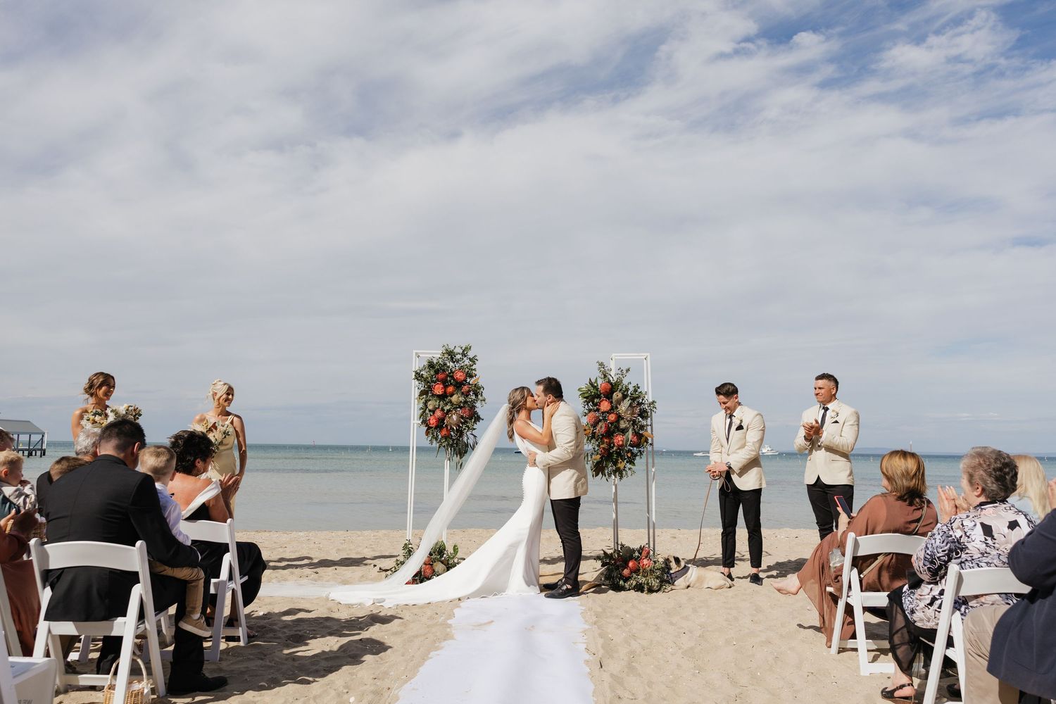 Couple shares first kiss at romantic beach wedding ceremony with floral arch, guests seated on white chairs by the ocean.